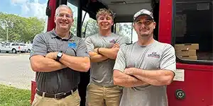 three adults standing at entrance of double decker bus smiling with their arms crossed
