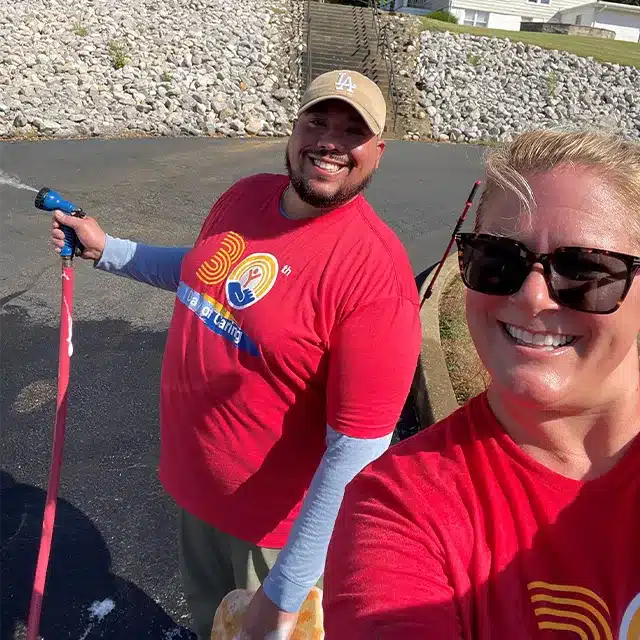 two adults wearing red shirts smiling at the camera whiling washing a bus